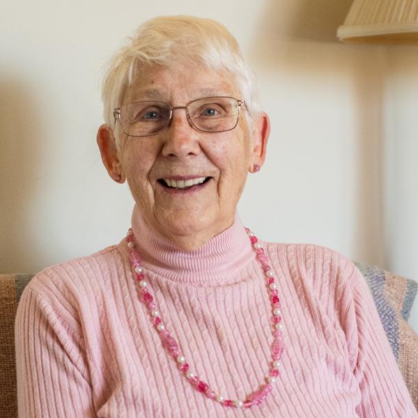 A person with short white hair and glasses is smiling warmly at the camera. They are wearing a light pink cable-knit sweater and a pink beaded necklace.